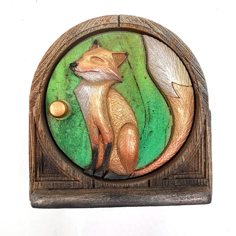 K&ouml;nnte beinhalten: Holzskulptur eines Fuchses mit detaillierter Schnitzerei. Der Fuchs ist orange und wei&szlig;, vor einem gr&uuml;nen Hintergrund in einem braunen, bogenf&ouml;rmigen Rahmen. Ein goldfarbener Knopf befindet sich auf der linken Seite. Die Skulptur hat ein rustikales, handwerkliches Aussehen.