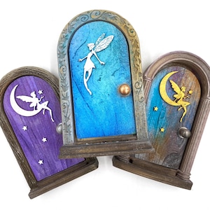May include: Three decorative fairy doors with arched frames. The doors are painted in shades of blue, purple, and brown, each featuring a fairy silhouette and a crescent moon or stars. The doors have small, round doorknobs.
