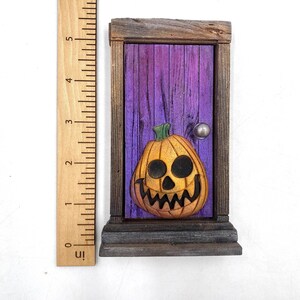 May include: A wooden Halloween-themed miniature door with a carved jack-o'-lantern. The door is painted purple with a silver doorknob, and the pumpkin is orange with black eyes and a jagged mouth. The door frame is made of weathered wood.