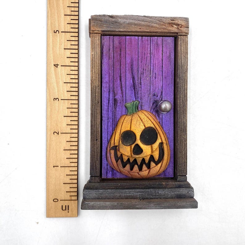 May include: A wooden Halloween-themed miniature door with a carved jack-o'-lantern. The door is painted purple with a silver doorknob, and the pumpkin is orange with black eyes and a jagged mouth. The door frame is made of weathered wood.