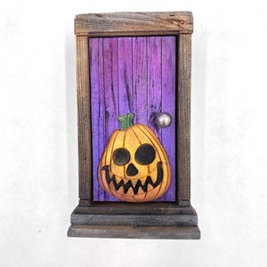 May include: A Halloween-themed wooden door decoration. The door is painted purple with a carved orange jack-o'-lantern. The door is framed in weathered wood, with a silver doorknob. The decoration has a small shelf at the bottom.