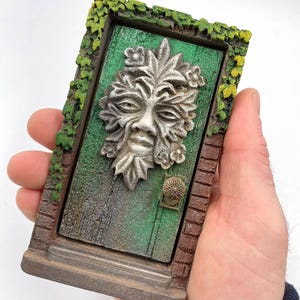 Puede incluir: Una puerta decorativa en miniatura con una superficie pintada de verde y una cara esculpida con detalles de hojas. La puerta tiene un pomo de color bronce y está enmarcada con ladrillos y vides verdes.