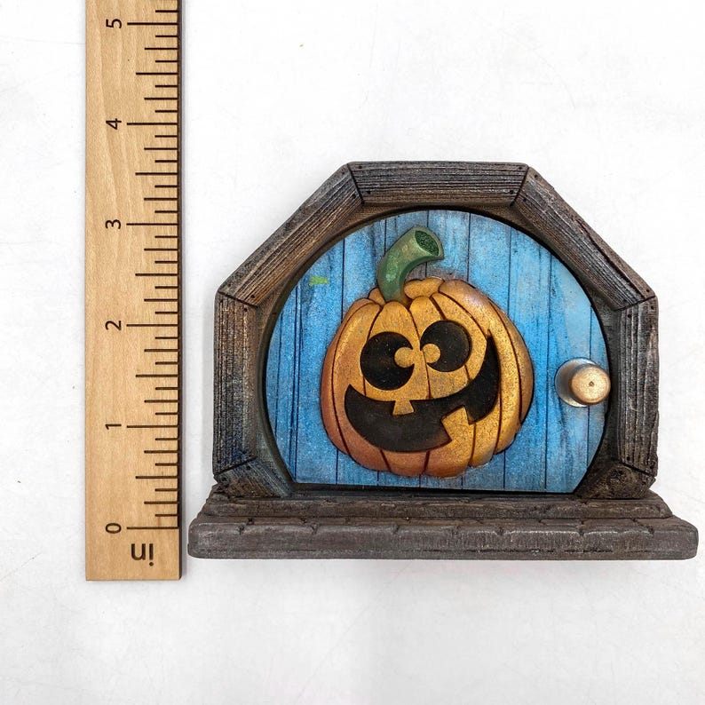 May include: A Halloween-themed fairy door with a carved pumpkin face on a blue door. The door has a wooden frame and a small brass doorknob. The door is approximately 5 inches tall.