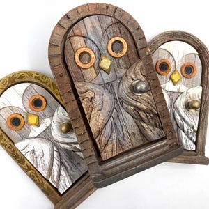 May include: Three wooden owl-shaped door decorations. Each features a carved owl face with circular eyes, a beak, and a door handle. The wood is stained in shades of brown, grey, and white, with gold and copper accents. The owl's eyes are dark brown.
