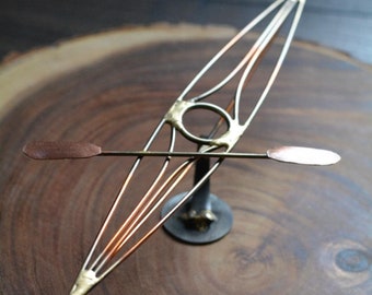 May include: A metal sculpture of a kayak with copper and gold accents, resting on a wooden base. The kayak is balanced on a thin metal rod with two copper paddles.