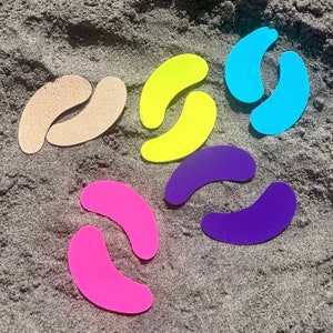 Op de afbeelding: Een assortiment kleurrijke, halvemaanvormige objecten in tinten beige, geel, lichtblauw, paars en roze, verspreid op een zanderige ondergrond. De vormen zijn glad en lijken van een vast materiaal te zijn gemaakt.
