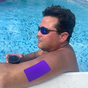 Op de afbeelding: Een persoon met een zonnebril ontspant in een zwembad en houdt een glas rode wijn vast. Een paarse rechthoekige pleister is op de arm van de persoon geplakt. Het zwembadwater is helderblauw.