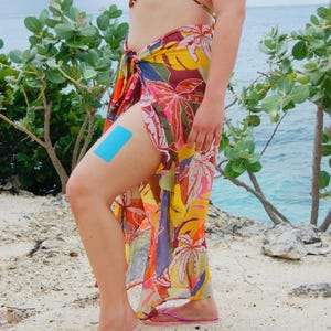 Op de afbeelding: Een kleurrijke sarong met een tropisch bloemenpatroon in tinten geel, oranje, roze en paars. Een blauwe kinesiotape is op de dij aangebracht. De persoon staat op een zandstrand met de oceaan op de achtergrond.