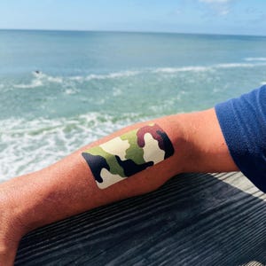 Op de afbeelding: Een kinesiotape met camouflagepatroon aangebracht op een arm. De tape heeft een mix van groene, bruine en zwarte vormen. De achtergrond toont de oceaan en een blauw shirt.