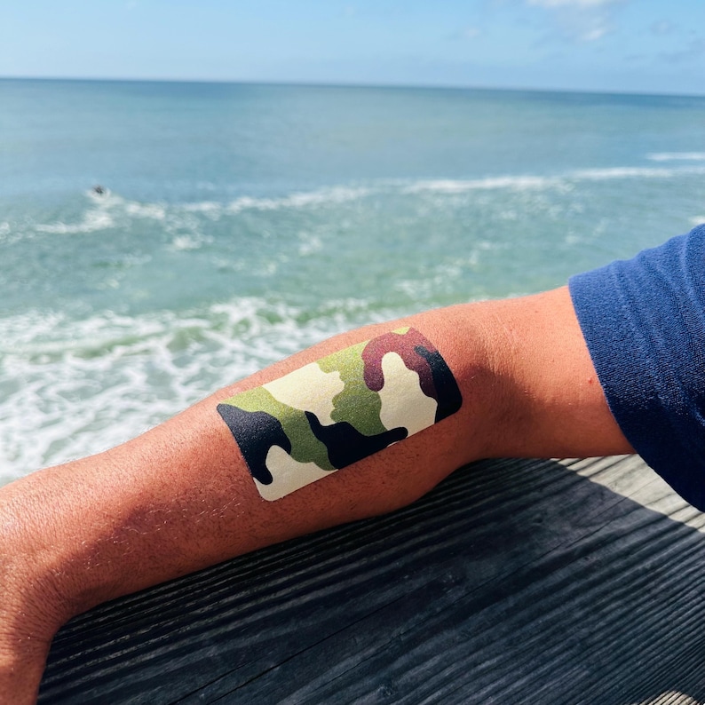 Op de afbeelding: Een kinesiotape met camouflagepatroon aangebracht op een arm. De tape heeft een mix van groene, bruine en zwarte vormen. De achtergrond toont de oceaan en een blauw shirt.