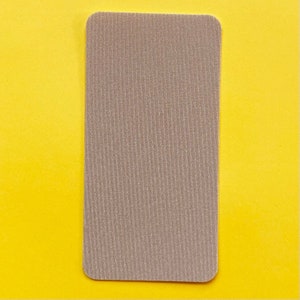 Op de afbeelding: Een rechthoekige, beige stoffen patch met afgeronde hoeken. De patch heeft een getextureerd, geribbeld oppervlak en staat tegen een heldergele achtergrond.