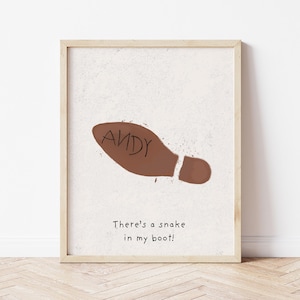 Puede incluir: Impresión artística enmarcada con una huella de bota marrón y el nombre "ANDY" en su interior. Debajo, el texto dice "There's a snake in my boot!". El fondo es blanco roto moteado y el marco es de madera clara.