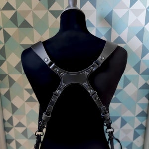 May include: A black leather dual camera harness with silver-toned rivets and adjustable straps. Two cameras are attached to the harness. The harness is displayed on a black mannequin against a geometric patterned wall.