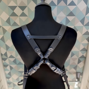 May include: A black leather camera harness with silver-toned metal hardware. The harness is designed to hold two cameras, with one camera attached on each side. The harness is displayed on a black mannequin.