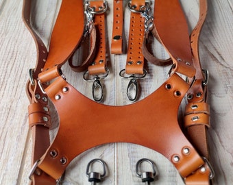 Può includere: Un cinturino per fotocamera in pelle marrone con cinturini regolabili e accessori in metallo. Il cinturino è progettato per essere indossato sul petto e ha una sezione larga e imbottita per il comfort.