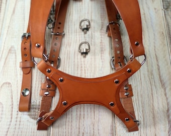 Può includere: Un cinturino per fotocamera in pelle marrone con cinturini regolabili e accessori in metallo. Il cinturino ha un pezzo per il petto e due bretelle.