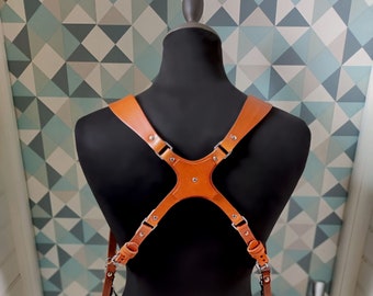 May include: A brown leather camera harness with two cameras attached. The harness is designed to be worn over the shoulders and across the back, with adjustable straps and metal hardware. The cameras are black with silver lenses.