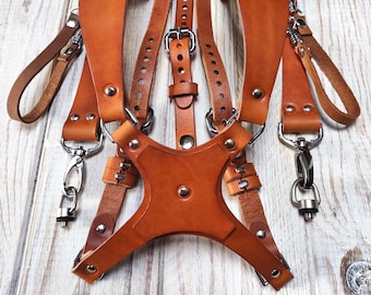 Può includere: Un cinturino per fotocamera in pelle marrone con più cinturini regolabili e fibbie metalliche. Il cinturino è progettato per essere indossato sul petto e sulle spalle.