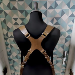 Peut inclure: Une sangle de caméra en cuir marron avec des accents métalliques et des sangles réglables. La sangle est portée sur un mannequin noir et est conçue pour tenir deux appareils photo.