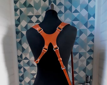 May include: A brown leather camera strap with silver hardware. The strap is designed to be worn across the chest and has adjustable straps. The strap is shown on a black mannequin.