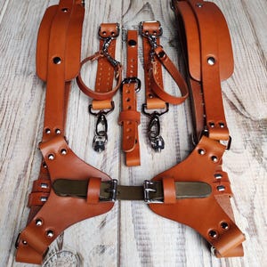 May include: A brown leather camera strap with two adjustable straps, two buckles, and two metal clips. The strap is designed to be worn across the chest and has a shoulder pad.