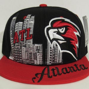 May include: Black and red baseball cap with a white and red Atlanta Falcons logo and a cityscape design. The cap has a red brim with the word "Atlanta" written in black.