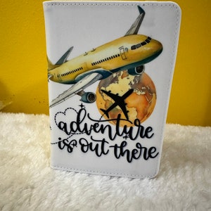 May include: White passport holder with a yellow airplane flying over a globe. The text "adventure is out there" is written in black cursive.