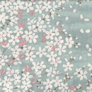 May include: A textile featuring a floral design. White cherry blossom flowers with pink details and falling petals are set against a pale teal backdrop. The pattern creates a feeling of spring and nature.