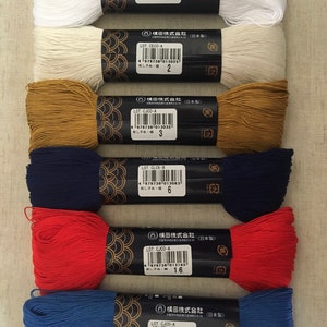 Könnte beinhalten: Sechs Spulen mit Stickgarn in verschiedenen Farben, darunter Weiß, Beige, Braun, Dunkelblau, Rot und Blau. Jede Spule hat ein Etikett mit dem Markennamen "Yokota", der Losnummer und der Farbnummer. Die Etiketten sind schwarz mit goldenen Akzenten.