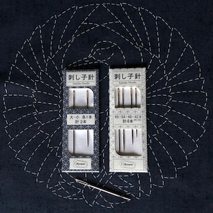 Könnte beinhalten: Zwei Packungen Sashiko-Nadeln mit japanischem Text und eine silberne Nadel auf dunkelblauem Stoff mit weißen Nähten. Die Packungen enthalten verschiedene Nadelgrößen. Der Stoff hat ein kreisförmiges Stichmuster.