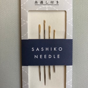 Könnte beinhalten: Eine Packung Sashiko-Nadeln mit goldfarbenen Ösen. Die Nadeln sind unterschiedlich lang und werden auf einer weißen Karte mit japanischem Text oben und dem Wort "DARUMA" unten präsentiert. Die Karte hat ein blaues Band mit den Worten "SASHIKO NEEDLE".
