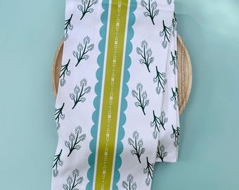 Puede incluir: Toalla de cocina blanca con un patrón repetido de ramitas verde oscuro. Una franja vertical de bordes festoneados turquesa y una franja central verde lima con detalles punteados. La toalla está doblada sobre un plato de madera.