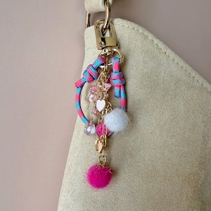 Puede incluir: Un llavero decorativo con un cierre dorado y un lazo de cuerda azul y rosa. El dije presenta un pompón blanco, un corazón rosa, cuentas rosas y un pompón fucsia. El llavero está sujeto a un bolso de ante beige.