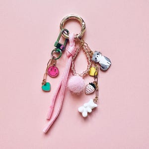 以下が含まれることがあります： ピンク色の背景に、ピンク色のコードと複数のチャームが付いたシルバーのキーホルダー。チャームには、笑顔、緑色のハート、ピンクのポンポン、イチゴ、白いクマ、黄色のベル、グレーと白の猫が含まれています。