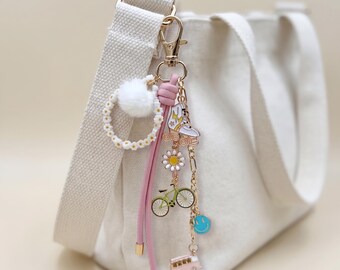 May include: A white canvas tote bag with a gold-colored clasp and a collection of colorful charms. The charms include a daisy, a bicycle, a bus, a thumbs-up, a sneaker, and a smiley face. A pink leather strap and a white pom-pom add to the bag's design.