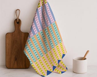 May include: A colorful kitchen towel with a geometric pattern of triangles and stripes in blue, pink, yellow, and green. The towel is hanging on a white wall next to a wooden cutting board and a small white cup with a wooden spoon.