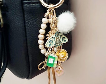 Op de afbeelding: Een goudkleurige sleutelhanger met meerdere bedels, waaronder een mot, een klein boek, een theekopje en een hart. De sleutelhanger heeft ook een streng parels en een pluizige witte pompon. De achtergrond is een zwarte tas.