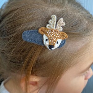 May include: A handcrafted hair clip with a deer motif. The clip features a grey felt base, with a deer head made from brown, white, and gold felt. The deer has white spots and embroidered details. Designed for use in the hair.