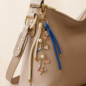 May include: A beige handbag with a gold-tone clasp and strap. A charm hangs from the clasp, featuring a white cord, blue cord, and gold chain with seashell, starfish, and pearl charms. The charms are in pink, blue, and gold.