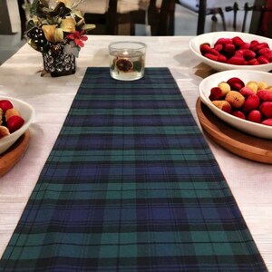 May include: A green, blue, and black plaid table runner with a white background. The runner is on a white table with a clear glass candle holder and a bowl of red and yellow fruit.