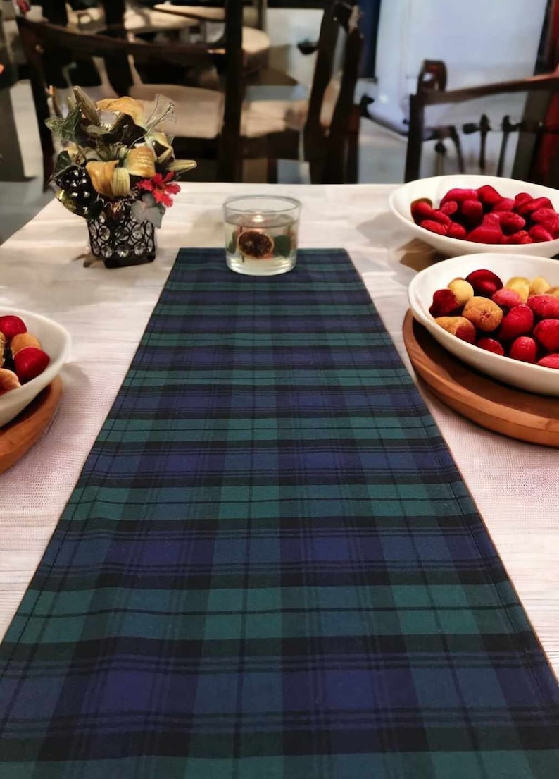 May include: A green, blue, and black plaid table runner with a white background. The runner is on a white table with a clear glass candle holder and a bowl of red and yellow fruit.