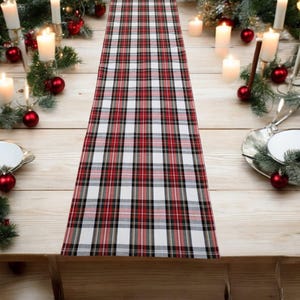 Peut inclure: Une nappe à carreaux rouge, noir et blanc. Le chemin de table est posé sur une table en bois clair, entouré de bougies, de boules rouges et de verdure, créant une ambiance festive.