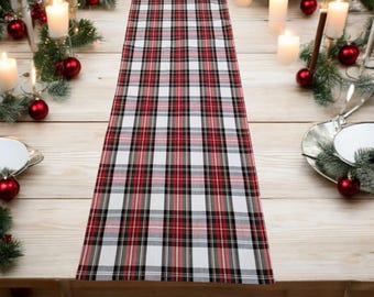 May include: A red, black, and white plaid table runner. The runner is centered on a light wood table, surrounded by candles, red ornaments, and greenery, creating a festive holiday setting.