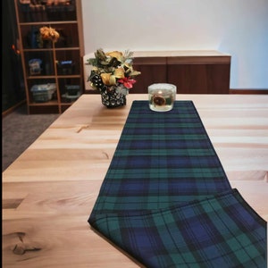 May include: A green and blue plaid table runner with a candle in a glass jar on top of it. The table runner is on a light wood table.
