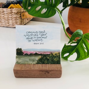 May include: A wooden stand with a white card featuring a watercolor painting of a landscape with a quote from Psalm 37:5, "Commit your way to the Lord, trust in Him and He will act."