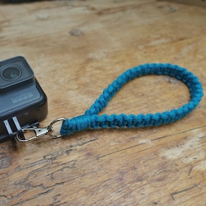 May include: A black GoPro camera with a teal braided wrist strap attached to the side.