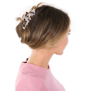 Puede incluir: Una pinza para el cabello con base blanca y un patr&oacute;n de carey en marr&oacute;n oscuro y negro. La pinza est&aacute; dise&ntilde;ada con dientes entrelazados y un mecanismo de resorte, sujetando una secci&oacute;n de cabello en un estilo recogido hacia arriba.
