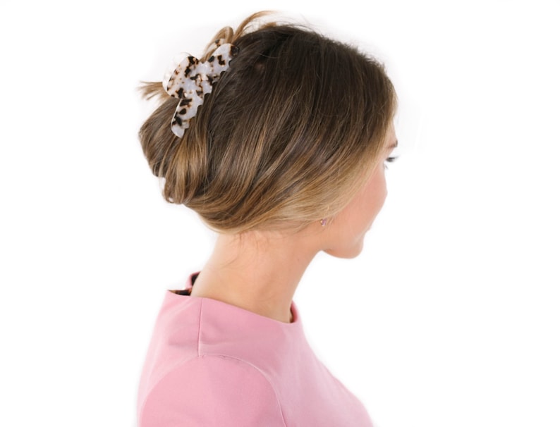 Puede incluir: Una pinza para el cabello con base blanca y un patr&oacute;n de carey en marr&oacute;n oscuro y negro. La pinza est&aacute; dise&ntilde;ada con dientes entrelazados y un mecanismo de resorte, sujetando una secci&oacute;n de cabello en un estilo recogido hacia arriba.
