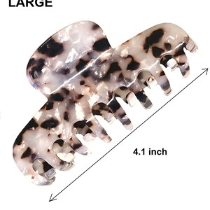 Puede incluir: Una pinza de pelo grande con un patr&oacute;n de carey blanco y negro. La pinza presenta una serie de dientes entrelazados y est&aacute; etiquetada como LARGE con una medida de 10,4 cm indicada por una flecha de doble punta en la imagen.
