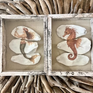 May include: Two framed seashell art pieces. One features a mermaid with a floral crown and flowing hair, holding a sprig. The other displays a seahorse. Both are painted in shades of brown and beige, set against a neutral background, and framed in white.